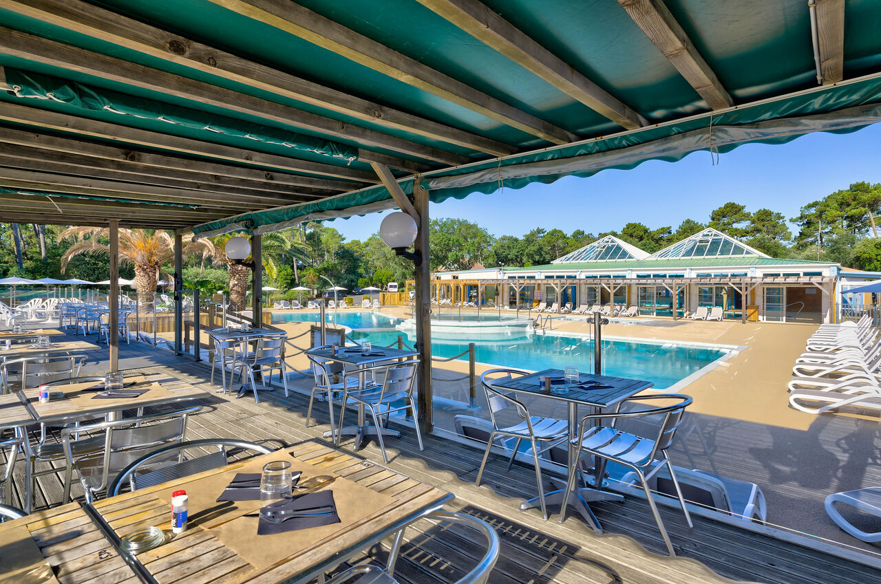 Terraza de restaurante sombreada con vista a la gran piscina exterior en el camping LIBRANOO Naturiste Arnaoutchot en Veille-Saint-Girons, Landes, Francia.