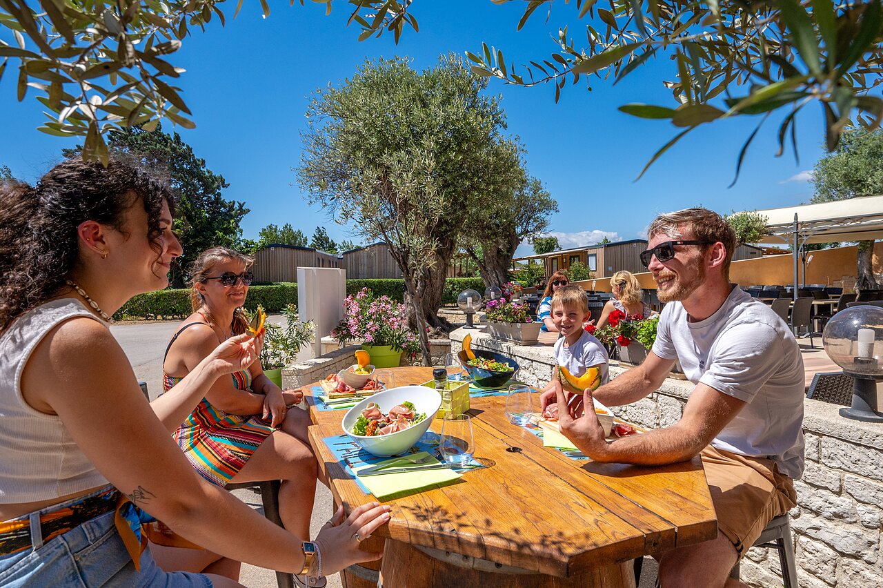 Familia sonriente almorzando en terraza sombreada, mobil-homes en camping CAPFUN Barbacane en NARBONNE (11).