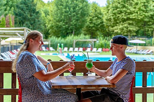 Pareja sonriente con cócteles en el bar de la piscina, en el camping CLICOCHIC Beau Rivage en LA ROQUE-GAGEAC (24).
