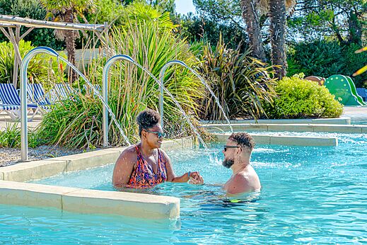 Piscina, chorros de agua y pareja sonriente en el camping VAGUES OCEANES Blancs Chênes en LA TRANCHE SUR MER (85).
