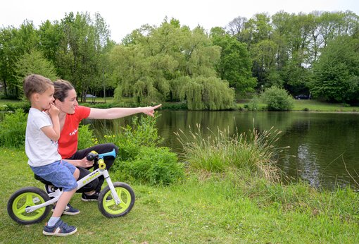 Niño en bicicleta de equilibrio y mujer señalando el lago en camping CAPFUN Breteche en Les Epesses (85).