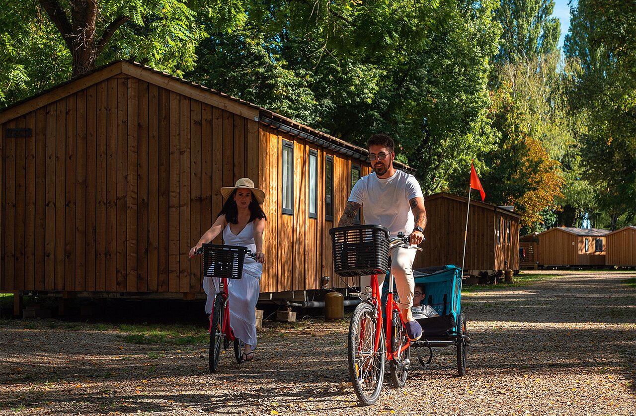 Pareja en bicicleta con remolque infantil frente a alojamientos en camping CLICOCHIC Au Clair Ruisseau en Gerstheim (67).