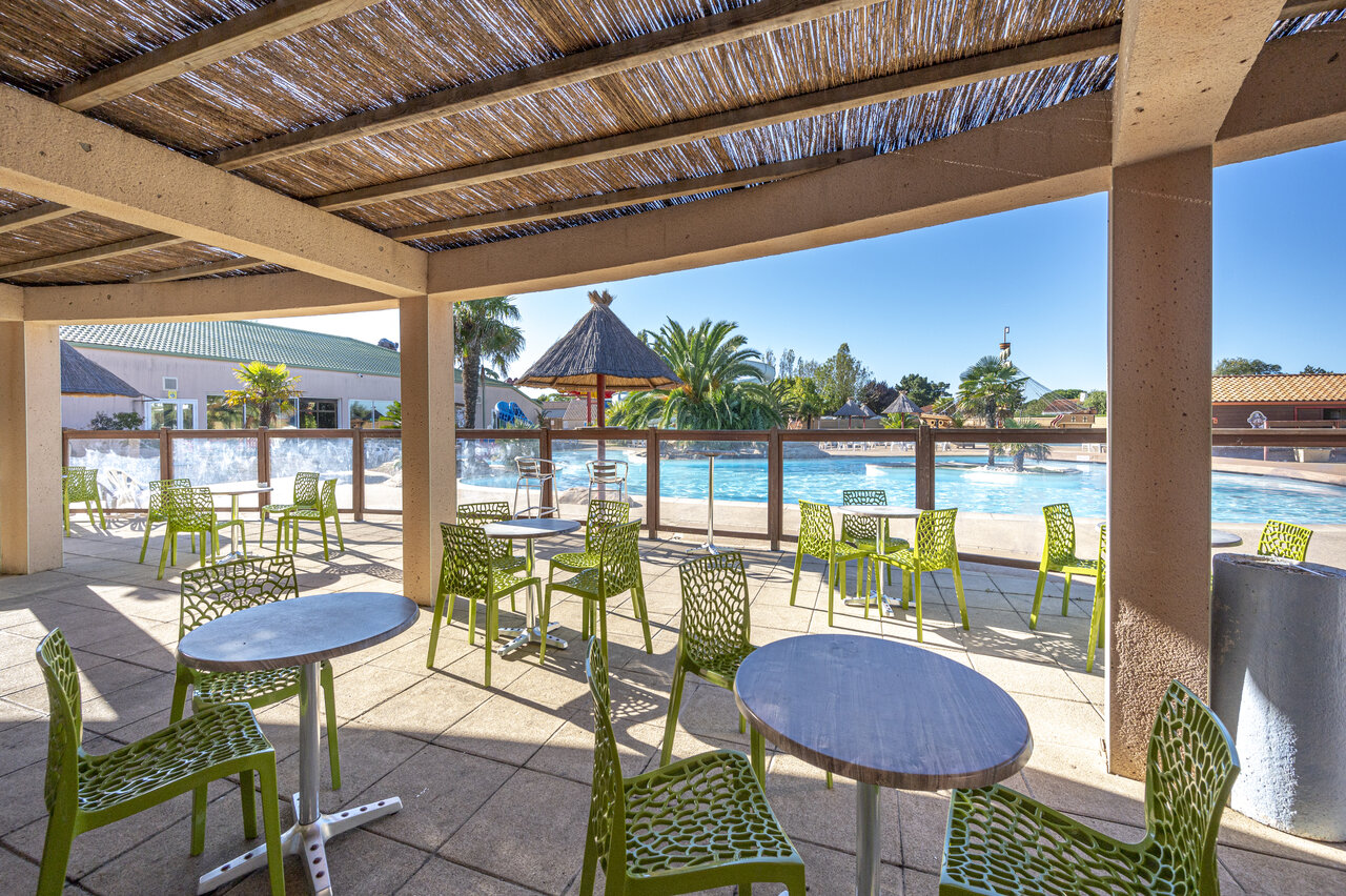 Terraza sombreada del bar con mesas y sillas verdes, junto a la piscina, en el camping CAPFUN Dauphins Bleus en GIVRAND (85).