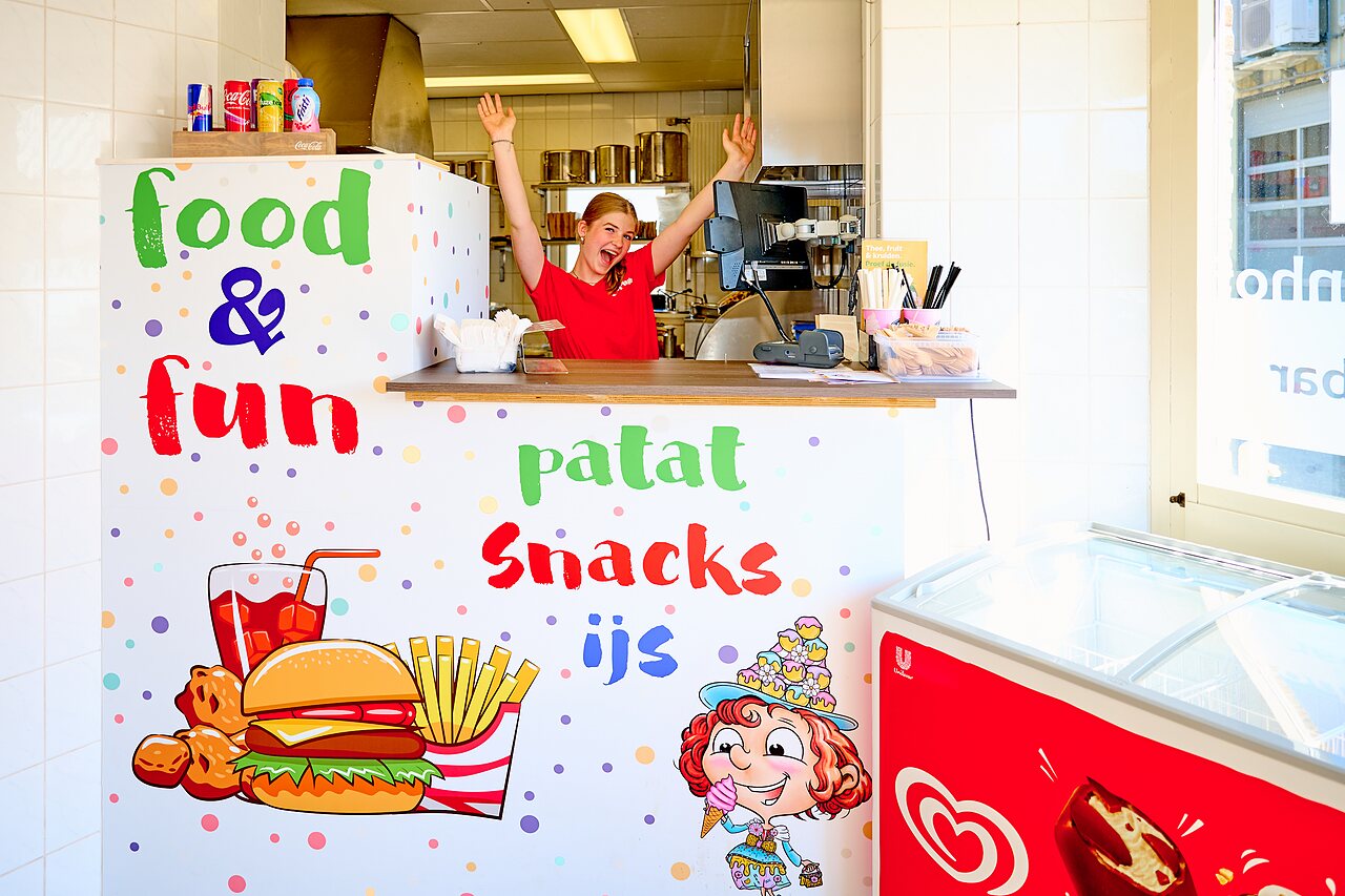 Mostrador de snack bar colorido con empleada sonriente, patatas fritas y helados en el camping CAPFUN De Eikenhof en Paasloo/Oldemarkt.