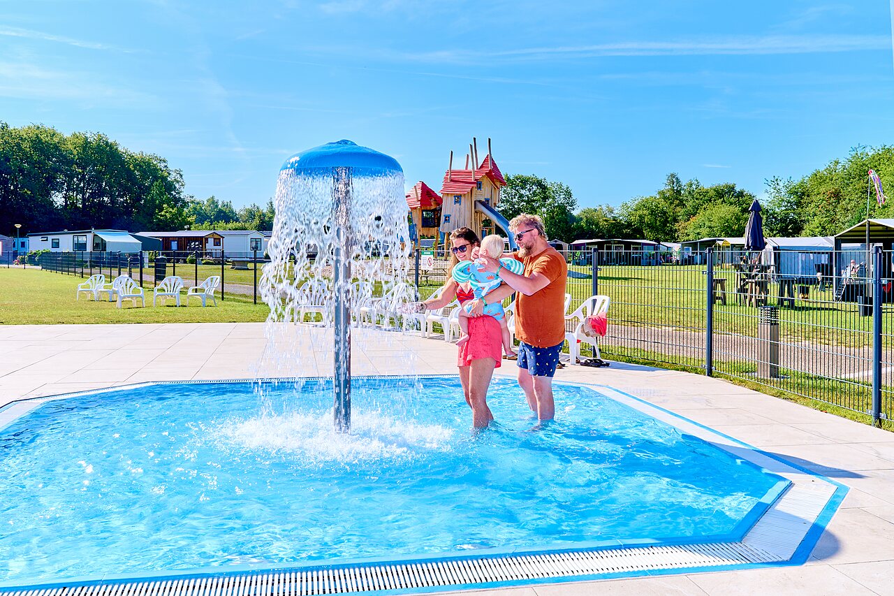 Familia con beb� en piscina infantil con fuente, parque infantil en camping CAPFUN De Eikenhof en Paasloo/Oldemarkt.