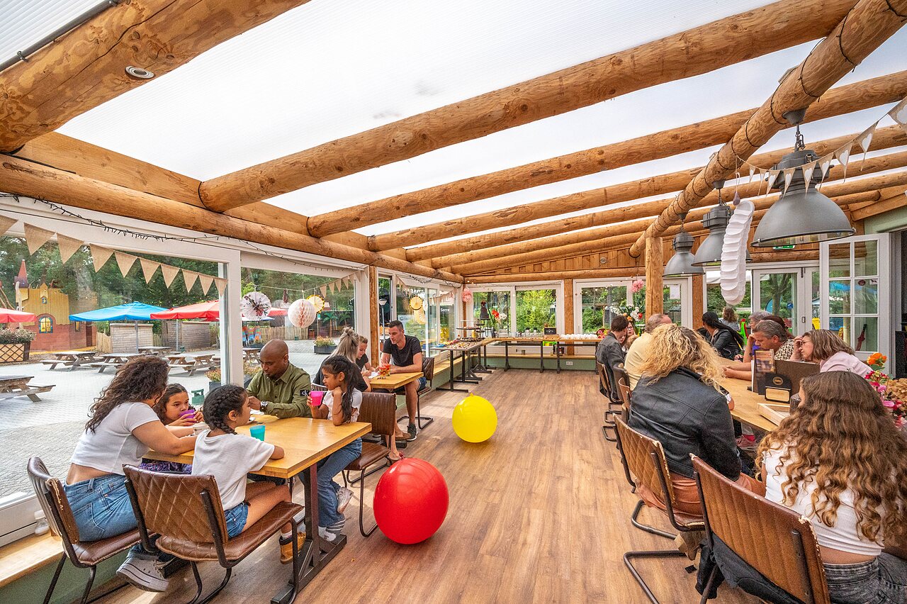 Familias y ni�os cenando en restaurante cubierto con vista al �rea de juegos en el camping CAPFUN De Scheepsbel en Doornspijk.
