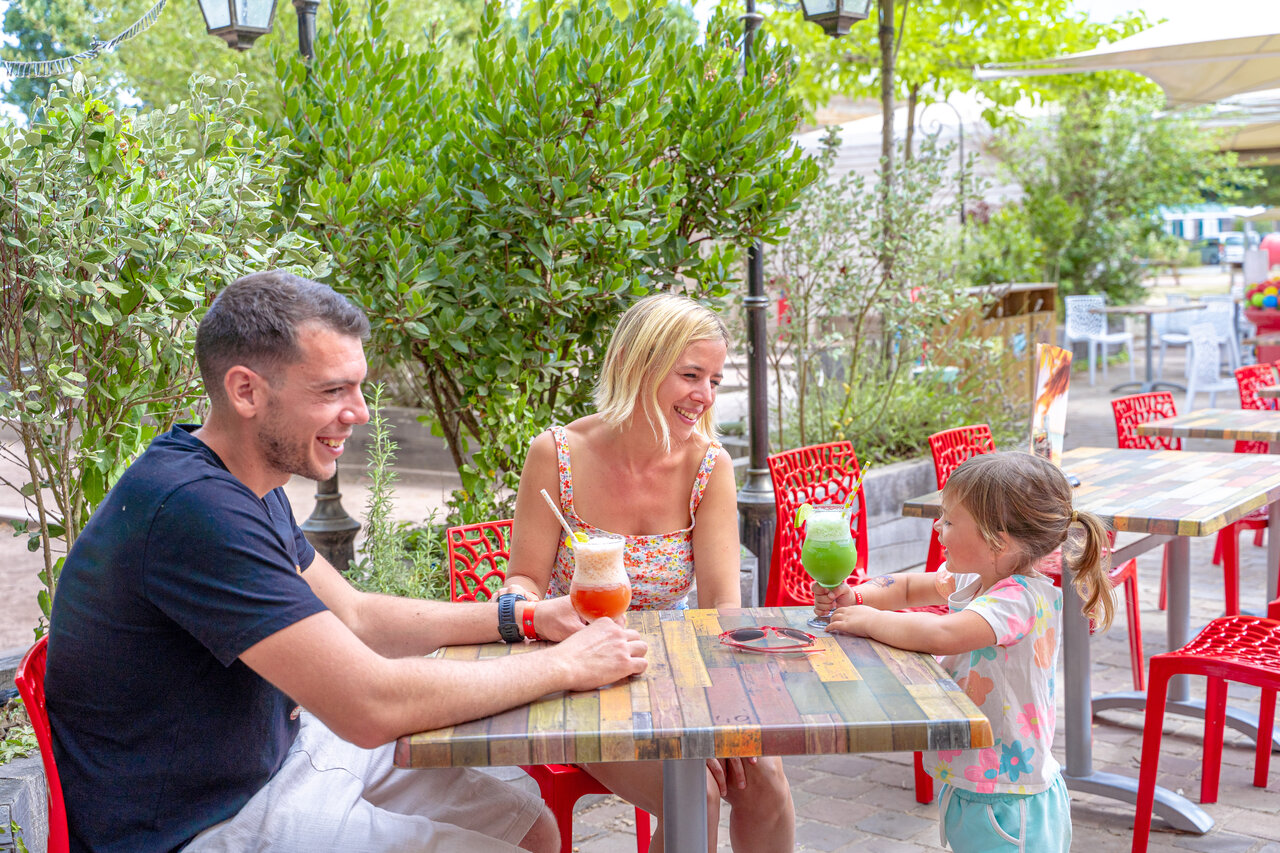 Familia sonriente disfrutando bebidas en terraza del restaurante, camping CAPFUN Donjon de Lars.