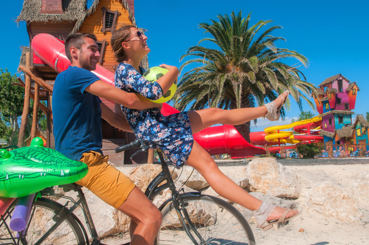Pareja feliz en bicicleta frente a toboganes acu�ticos coloridos en el camping CAPFUN F�erix en MARSEILLAN PLAGE (34)