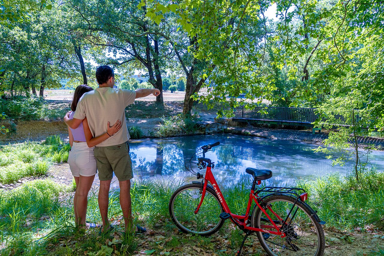 Pareja y bicicleta roja junto a un estanque arbolado en el camping CAPFUN Fumades en ALLEGRE-LES-FUMADES (30).