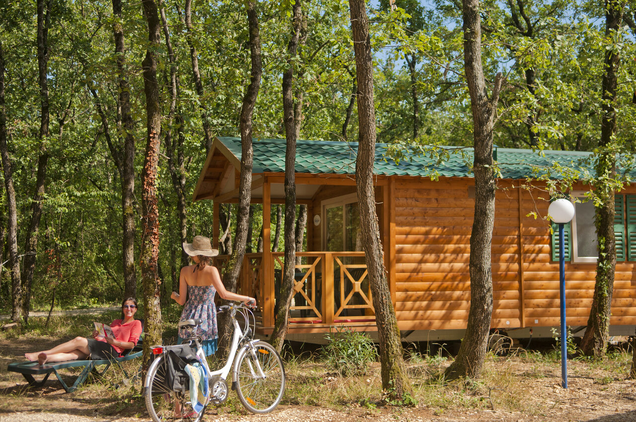 Chalet de madera, mujer en bicicleta y otra leyendo, bajo los �rboles en el camping CAPFUN Grand Lierne en CHATEAUDOUBLE (26).