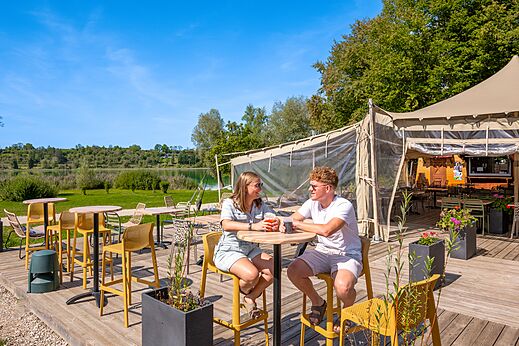 Terraza del bar-restaurante con pareja y lago en el camping CLICOCHIC Grisière.