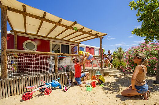 Mobil-home rojo con terraza, familia y niños jugando en el camping CAPFUN Hermitage en SERIGNAN (34).