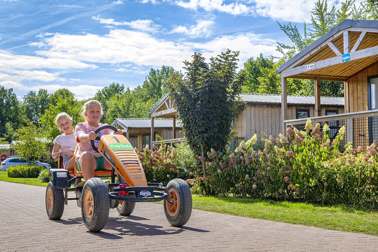 Ni�os sonrientes en kart a pedales frente a chalets en el camping CAPFUN IJsselstrand en Doesburg.