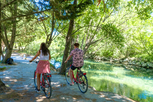 Pareja en bicicleta junto a un río sombreado en el camping CAPFUN Jantou en LE THOR (84).