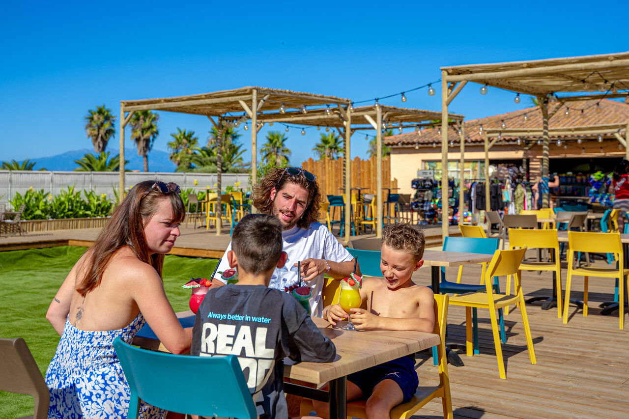 Familia sonriente disfrutando bebidas en la terraza del bar en el camping CAPFUN Las Bousigues en Le Barcar�s (66).