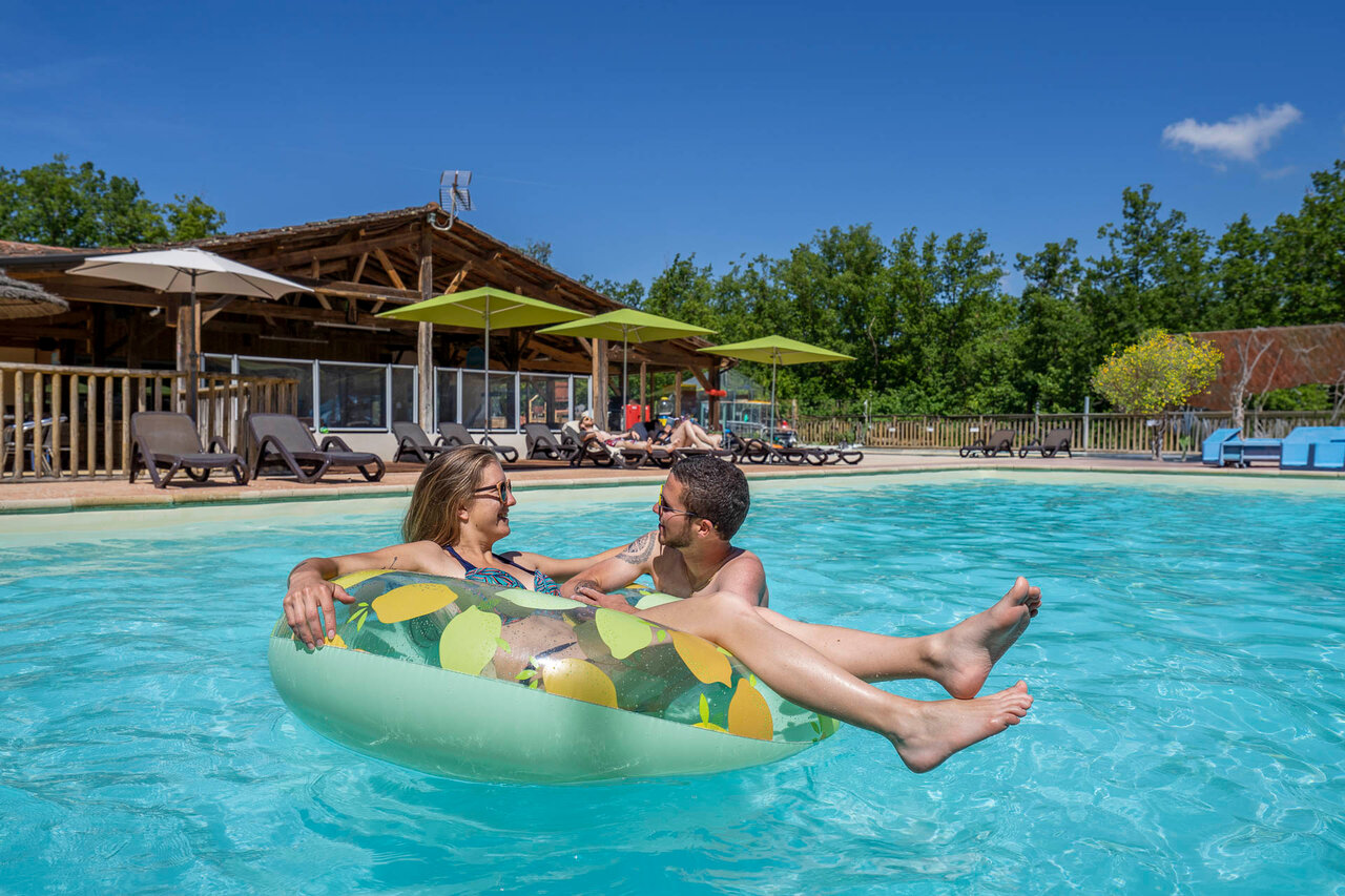 Piscina exterior, pareja sonriente en flotador. Camping CLICOCHIC Lou Castel.