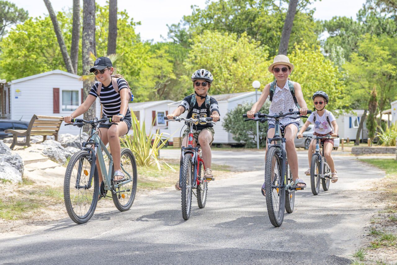 Ni�os sonrientes en bicicleta por un camino, Mobil-homes al fondo en el camping VAGUES OCEANES Marina Landes en Mimizan (40).