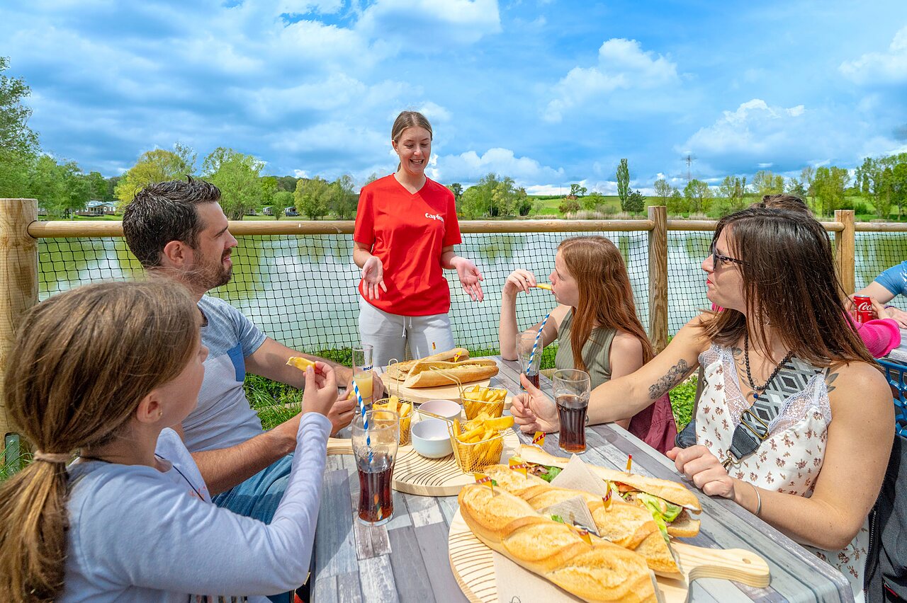 Familia almorzando en terraza con vista lago, en el camping CAPFUN Mirabelle en VOLSTROFF (57).