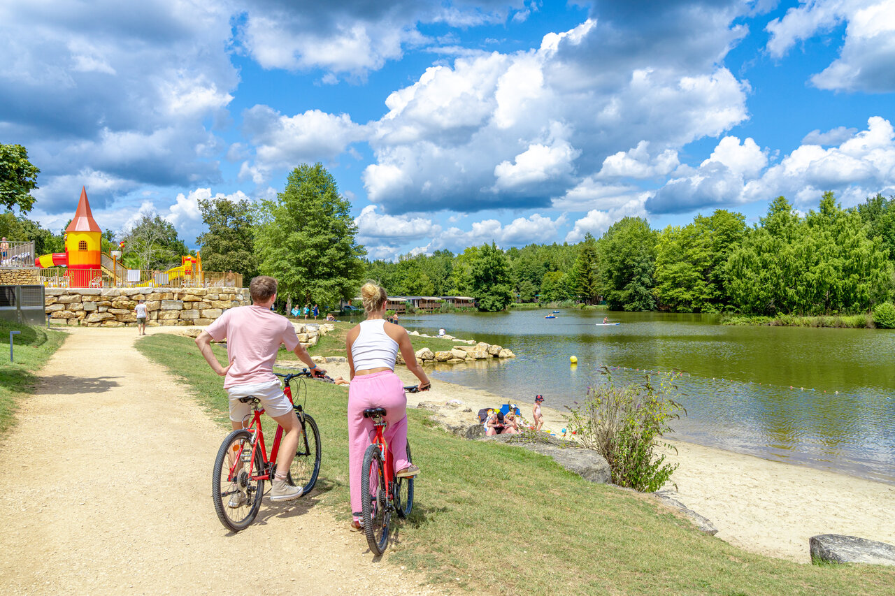 Pareja en bicicleta por sendero junto al lago y zona de juegos en camping CAPFUN Moulinal en BIRON (24).