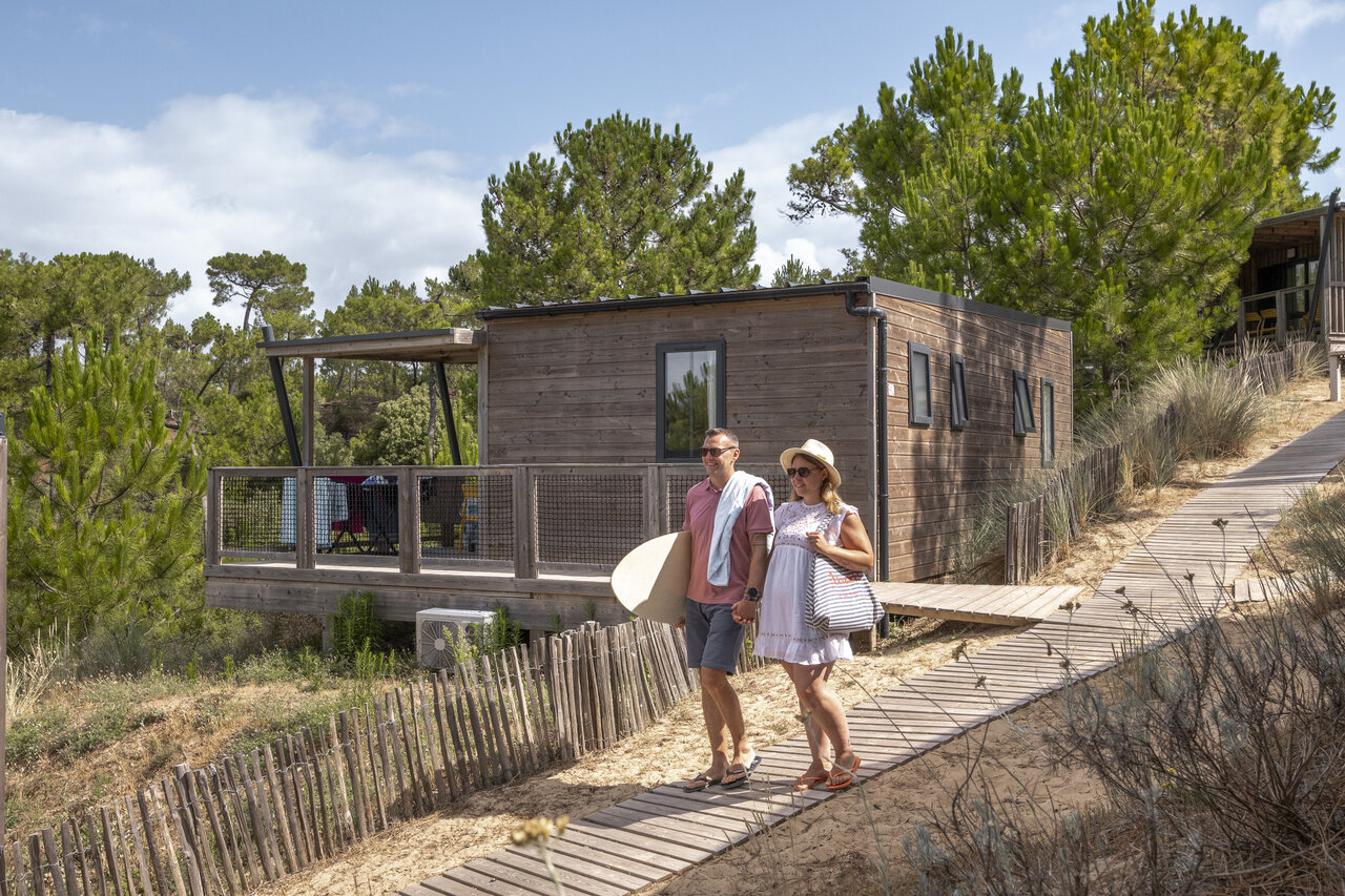Mobil-home moderno de madera con terraza, pareja y bodyboard en el camping CLICOCHIC Petit Bec en St Hilaire de Riez (85).