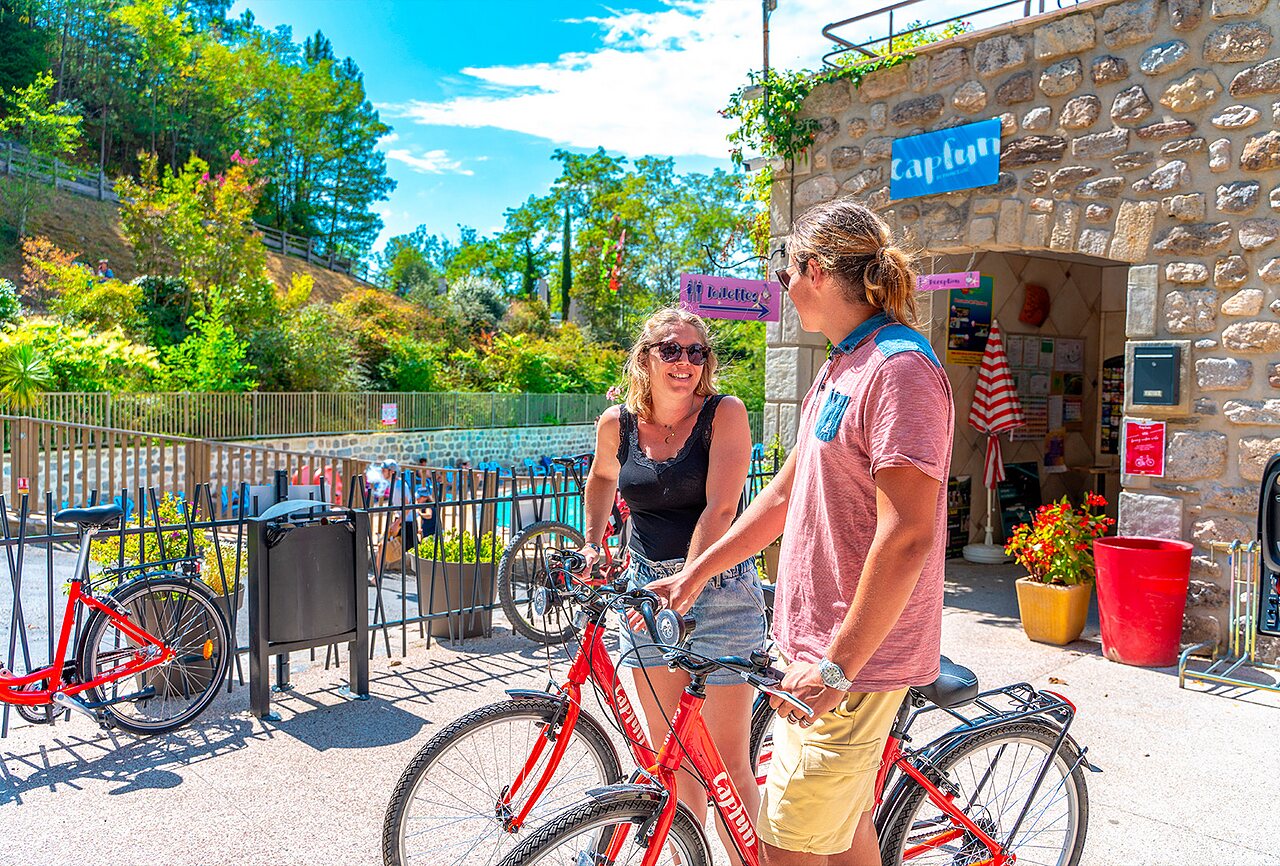 Dos personas sonrientes con bicicletas rojas cerca de la piscina en el camping CAPFUN Plantas en SAINT VINCENT DE DURFORT (07).