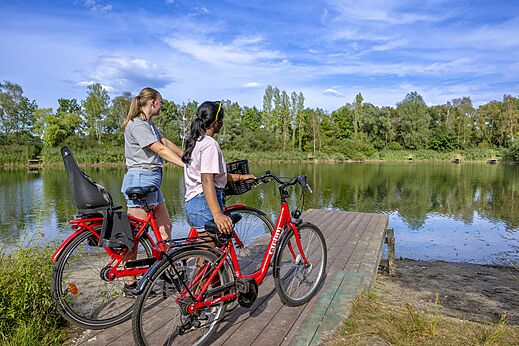 Bicicletas rojas y lago natural con dos personas en el camping CAPFUN Rakelbos.