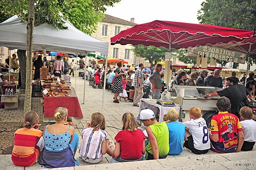 Mercado local animado en una plaza de pueblo, con puestos y visitantes en Lot-et-Garonne.