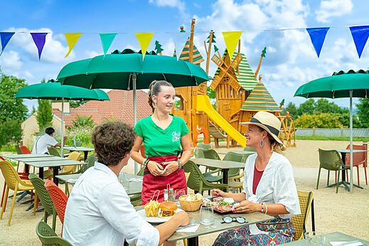 Camarera sirviendo a clientes en restaurante exterior con parque infantil en camping CLICOCHIC Village des Meuniers en Dompierre-les-Ormes (71).