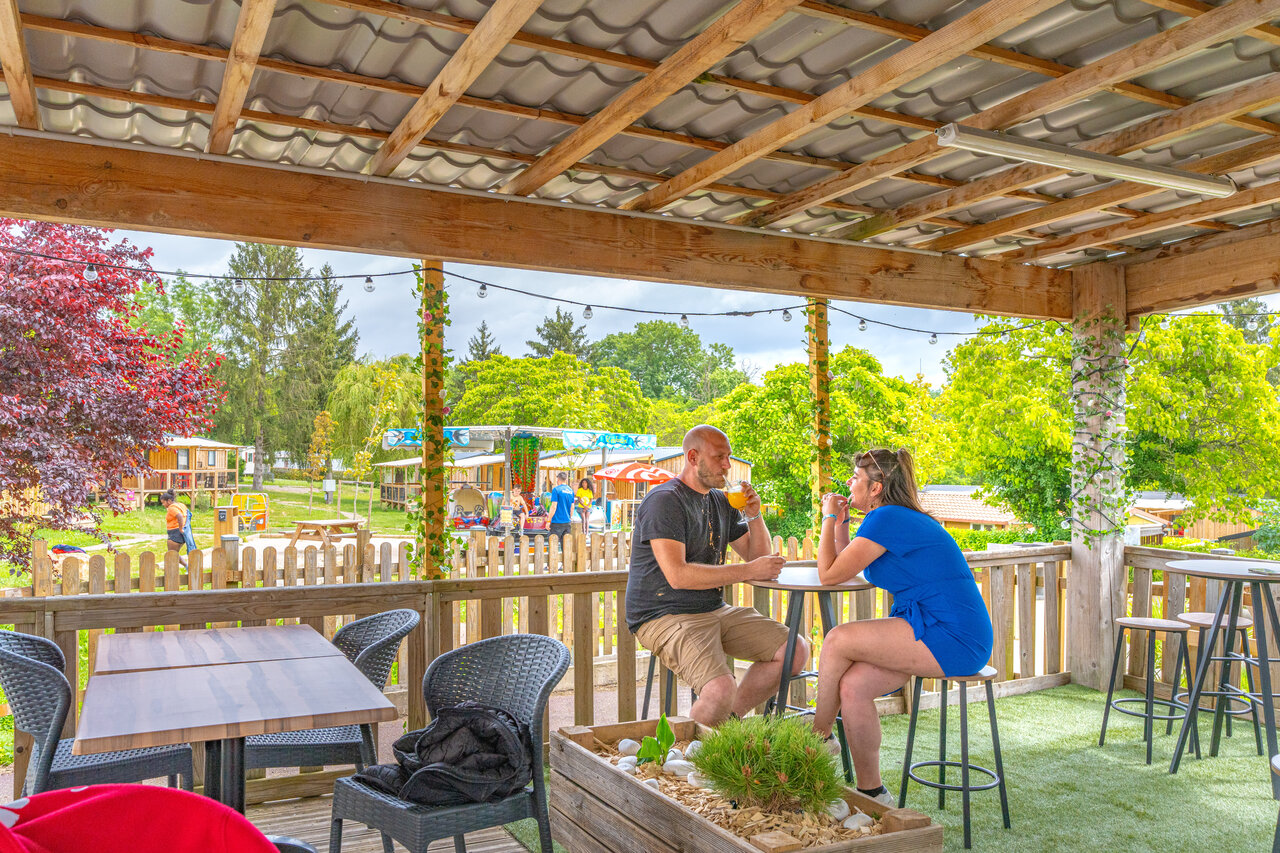 Terraza de bar animada, pareja y zona de juegos en el camping CAPFUN Le Village Parisien en VARREDDES (77).